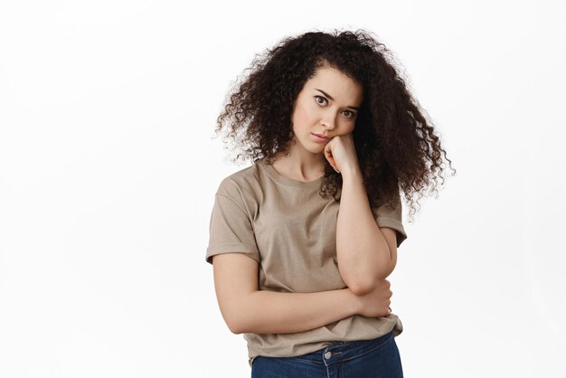 Image d'une femme brune réticente, tête penchée sur la main et regardant indifférente à la caméra, se sentant ennuyée, fatiguée d'écouter debout sur fond blanc