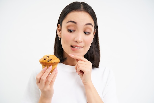 Photo gratuite image d'une femme coréenne pensive regarde un cupcake avec un visage pensif en pensant au dessert bac blanc