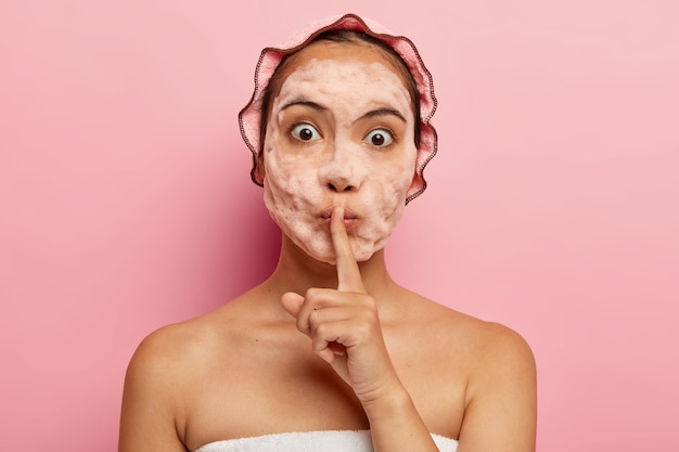 Image d'une femme coréenne surprise avec des bulles de savon sur le visage, fait un geste de silence, raconte un secret de beauté, nettoie et exfolie la peau, a des procédures cosmétiques pendant le temps libre, prend soin d'elle-même