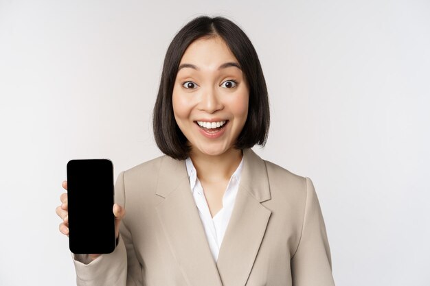 Image d'une femme d'entreprise asiatique montrant l'écran du téléphone mobile de l'interface de l'application faisant l'expression du visage surpris wow debout sur fond blanc