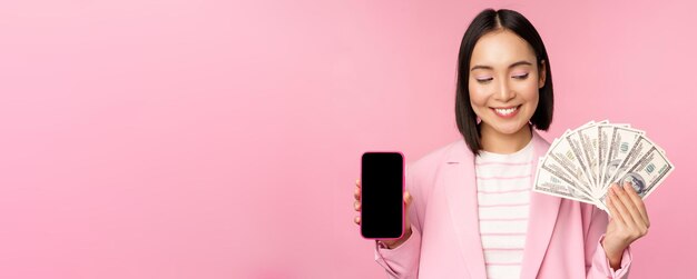 Image d'une femme d'entreprise coréenne prospère montrant des dollars d'argent et une interface d'écran d'application pour smartphone du concept d'application de téléphone mobile d'investissement et de financement fond rose