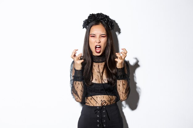 Image de femme folle en robe de dentelle gothique et couronne noire criant les poings en colère et serrant, debout en costume d'halloween avec une expression furieuse, debout sur fond blanc.