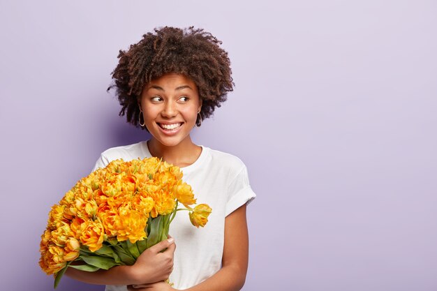 Image d'une femme joyeux anniversaire, célèbre une journée spéciale, obtient un gros bouquet de fleurs orange, porte un t-shirt décontracté, se concentre de côté, a le sourire sur le visage, regarde de côté, rencontre des invités, porte un t-shirt décontracté