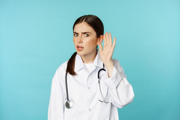Image d'une femme médecin confuse ne peut pas vous entendre, tenant la main près de l'oreille et ayant l'air perplexe, parlez un geste plus fort, fond de torquoise.