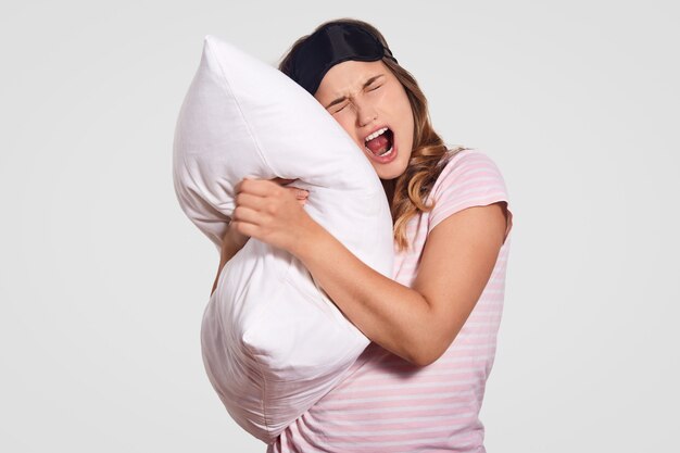 Image d'une femme qui bâille comme a une expression endormie, porte des vêtements domestiques, porte un oreiller blanc doux, pose sur blanc. Personnes, repos, confort, fatigue, concept de sommeil