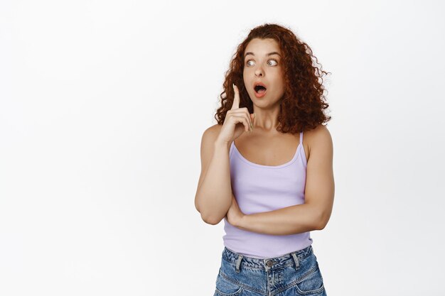 Image d'une femme rousse excitée qui lance une idée, lève le doigt et halete étonnée, a un plan, suggérant qch, a une solution, debout sur fond blanc