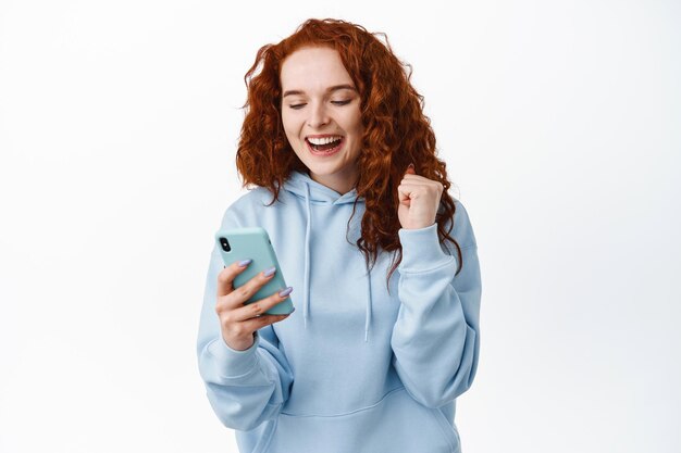 Image d'une femme rousse heureuse et satisfaite se réjouissant de regarder l'écran du smartphone, d'atteindre l'objectif de l'application, de lire de bonnes nouvelles, debout sur blanc