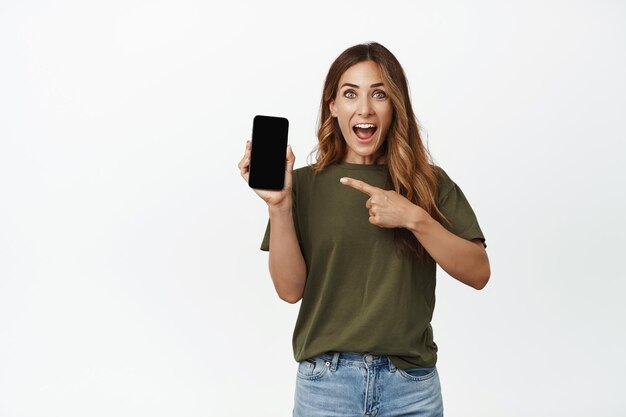 Image d'une femme souriante et heureuse présentant l'application, pointant du doigt l'écran du smartphone avec un visage étonné, démontrant une nouvelle fonctionnalité intéressante, une application d'achat en ligne, un arrière-plan blanc.