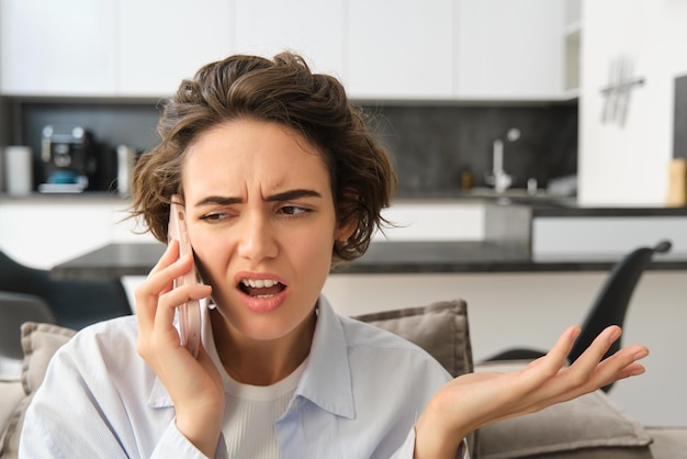 Image d'une femme avec un visage énervé et confus parlant au téléphone portable alors qu'elle est assise à la maison sur le canapé shru