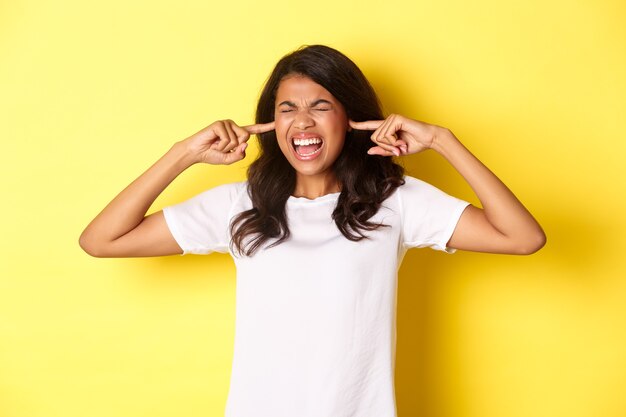 Image d'une fille afro-américaine énervée, ne peut pas supporter un bruit fort dérangeant, ferme les oreilles et crie agacé, debout sur fond jaune.