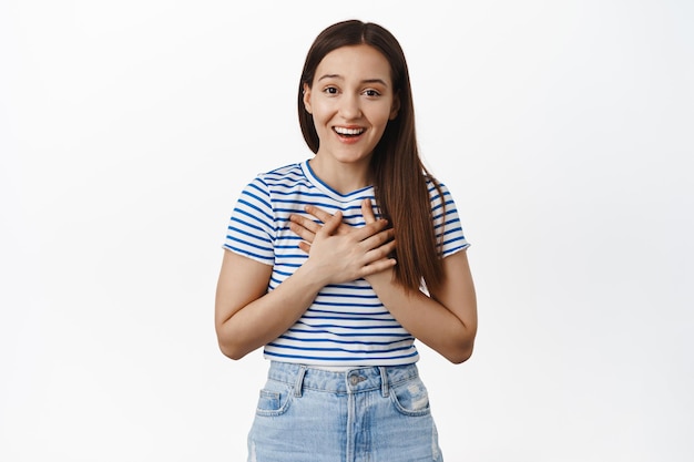 L'image de la fille a l'air touchée et flattée, tenant les mains sur le cœur et souriante reconnaissante, exprimant sa gratitude, merci de poser, debout sur fond blanc.