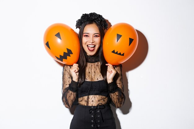 Image d'une fille asiatique en costume de sorcière maléfique tenant deux ballons orange avec des visages effrayants