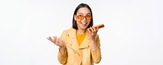Image d'une fille asiatique heureuse parlant sur un enregistrement sur haut-parleur traduisant sa voix avec une application de téléphonie mobile parlant dans un smartphone dynamique debout sur fond blanc