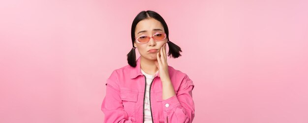 Image d'une fille asiatique triste boudant touchant sa joue faisant la moue déçue a mal aux dents debout sur fond rose