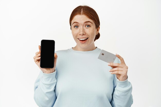 L'image d'une fille au gingembre souriante et excitée montre l'écran de son téléphone, son écran mobile vide et sa carte de crédit bancaire ou de réduction, debout dans des vêtements décontractés sur fond blanc.