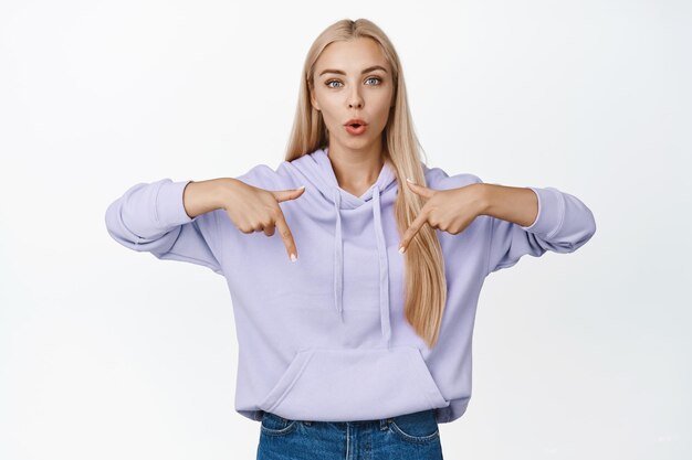 Image d'une fille blonde curieuse posant une question pointant les doigts vers le bas et regardant étonné debout sur fond blanc