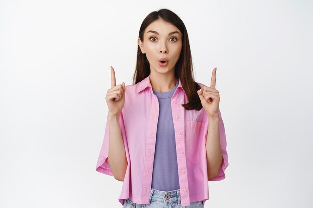 Image d'une fille brune surprise avec une expression de visage curieux pointant les doigts vers le haut intéressé par les remises de vente debout sur fond blanc