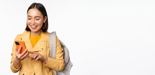 Image d'une fille heureuse touriste voyageur avec sac à dos regardant un smartphone à l'aide d'une application d'itinéraire de téléphone portable debout sur fond blanc Copier l'espace