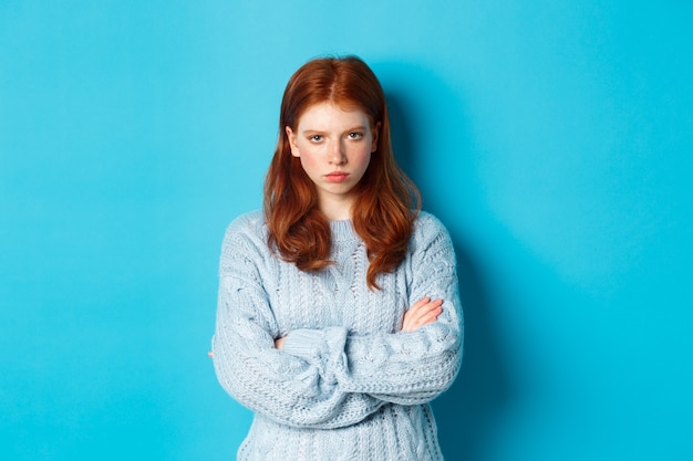 Image d'une fille rousse en colère se sentant offensée, les bras croisés sur la poitrine et boudant, regardant la caméra en colère, debout sur fond bleu.