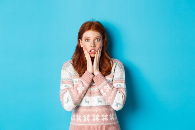 Image d'une fille rousse impressionnée haletant et regardant la caméra, entendez de grandes nouvelles, debout dans un pull d'hiver sur fond bleu.