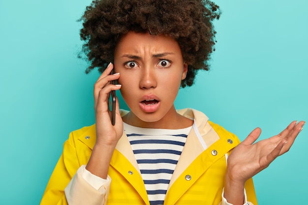 Image de gestes de femme afro-américaine frustrés avec paume pendant la conversation téléphonique, regarde avec indignation, vêtu d'un imperméable imperméable, isolé sur bleu, exprime l'aversion