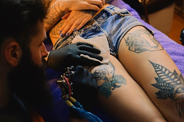 Image en gros plan de l'artiste tatoueur barbu fait un tatouage sur une jambe féminine.