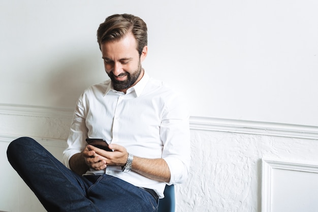 Image d'un homme d'affaires séduisant et heureux portant une chemise blanche en train de taper sur un téléphone portable et assis sur une chaise tout en travaillant au bureau