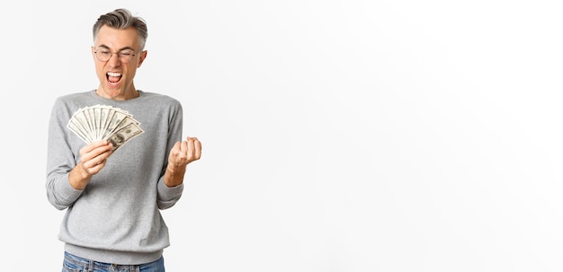 Image d'un homme d'âge moyen joyeux dans des verres et un pull gris gagnant de l'argent détenant de l'argent et se réjouissant