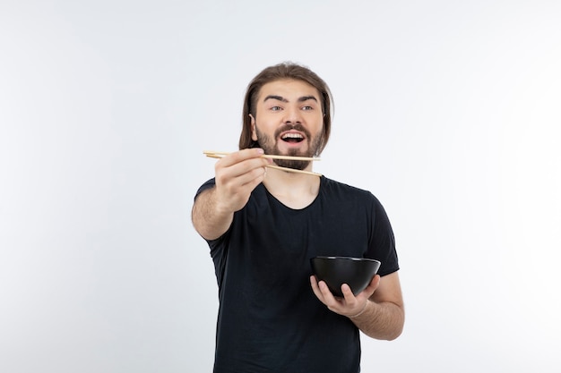 Image d'homme barbu tenant un bol avec des baguettes sur un mur blanc.