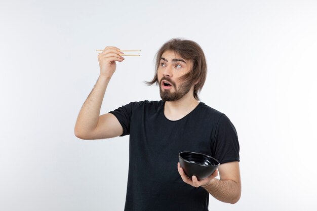 Image d'homme barbu tenant un bol avec des baguettes sur un mur blanc.