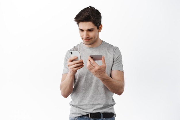 Image d'un homme brune regardant un smartphone tenant une carte de crédit en plastique transférer de l'argent avec une application payant dans les achats sur Internet debout sur fond blanc