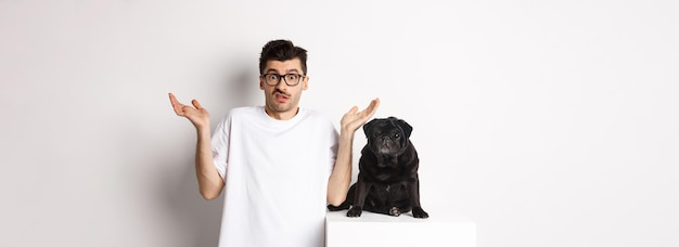 Photo gratuite image d'un homme confus dans des verres levant les mains et haussant les épaules compliqué debout près d'un chien carlin noir