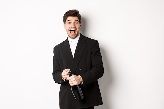 Image d'un homme séduisant en costume noir faisant une fête, célébrant le nouvel an et ouvrant une bouteille de champagne, debout heureux sur fond blanc