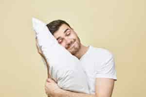 Photo gratuite image horizontale de beau jeune homme mignon avec des poils posant avec la tête sur un oreiller doux blanc dormant paisiblement et souriant, seeling bon rêve. beau mec sieste après une dure journée de travail