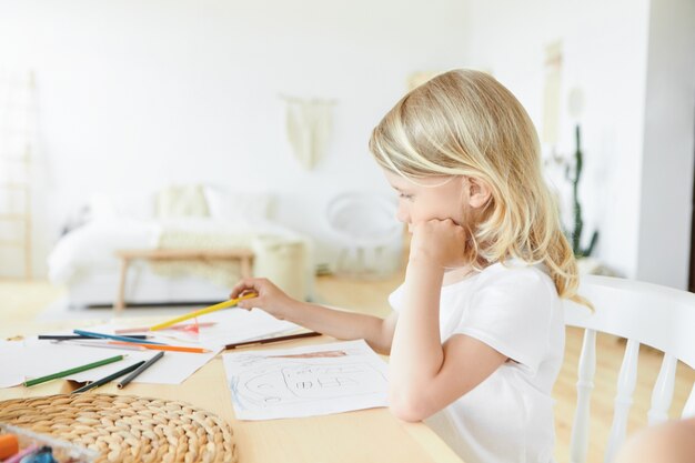 Image horizontale de petit enfant européen talentueux créatif avec des cheveux blonds lâches assis au bureau en bois dans l'intérieur de la chambre élégante avec des feuilles de papier et des crayons colorés, des traînées et de la peinture