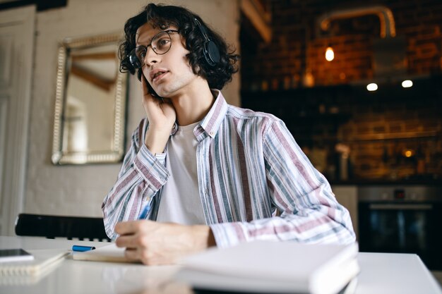 Image intérieure d'un jeune homme sérieux aux cheveux bouclés assis sur son lieu de travail avec des manuels, écrivant tout en écoutant des conférences via des écouteurs sans fil, en apprenant à la maison. Distanciation sociale