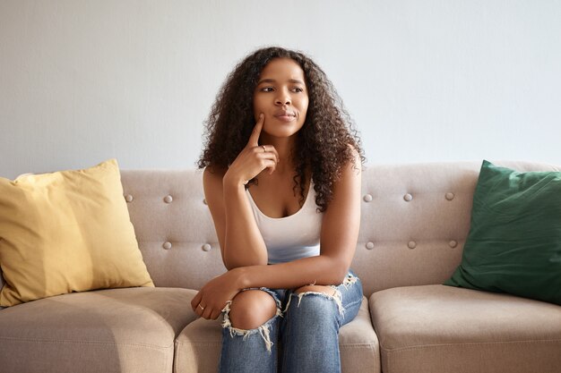 Image intérieure d'une jolie fille à la mode avec une apparence africaine assise isolée sur un canapé gris avec des coussins jaunes et verts ayant un regard réfléchi douteux, touchant le menton, disant Hmm, laissez-moi réfléchir