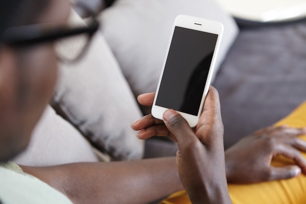 Image intérieure recadrée d'un homme à la peau sombre méconnaissable se détendre sur le canapé dans le salon, à l'aide du Wi-Fi à domicile sur un téléphone mobile moderne