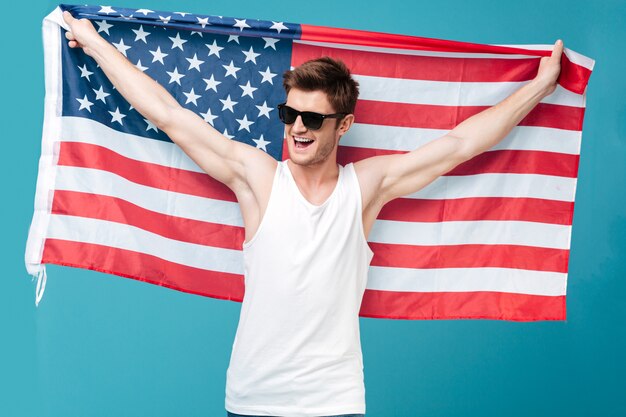 Image de jeune bel homme debout sur bleu isolé tenant le drapeau des États-Unis. En regardant de côté.