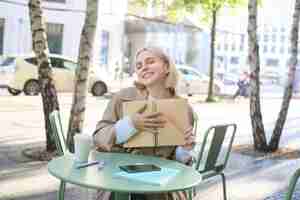 Photo gratuite image d'une jeune et belle étudiante faisant ses devoirs dans un café en plein air tenant un journal ou