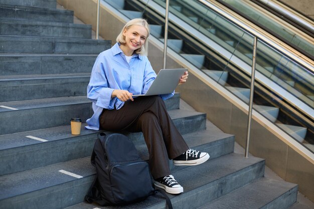 Photo gratuite image d'une jeune étudiante étudiant en ligne travaillant à distance assise sur les escaliers avec un ordinateur portable et