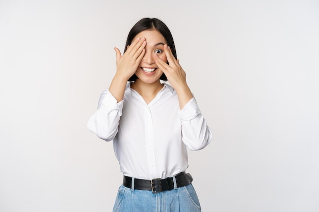 Image d'une jeune femme asiatique ferme les yeux et sourit attend la surprise anticipe jeter un coup d'œil à travers les doigts se dresse sur fond blanc