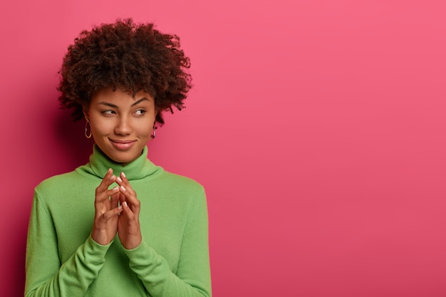 Photo gratuite image de jeune femme aux cheveux bouclés gestes avec intention, concentrée de côté, évènement intéressant, vêtue d'un col roulé vert, a une expression sournoise, modèles sur un mur rose, copiez l'espace de côté