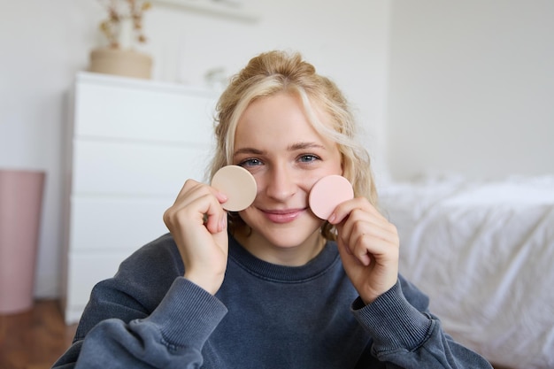 Photo gratuite image d'une jeune femme blonde qui enregistre une vidéo sur le maquillage, montre le ton de la peau, les produits de beauté, s'assoit dans un