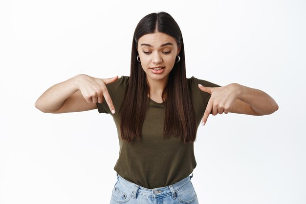 Image d'une jeune femme brune maladroite se mordant la lèvre et levant les sourcils tout en regardant et pointant vers le bas quelque chose d'étrange goutte sur fond blanc au sol