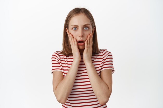 Image d'une jeune femme choquée et inquiète, haletant, regardant la caméra sans voix surprise, l'air inquiète, entendant de mauvaises nouvelles, debout sur fond blanc.