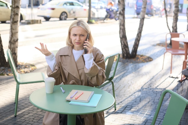 Image d'une jeune femme contrariée et confuse qui parle au téléphone portable et hausse les épaules assise dans un café.