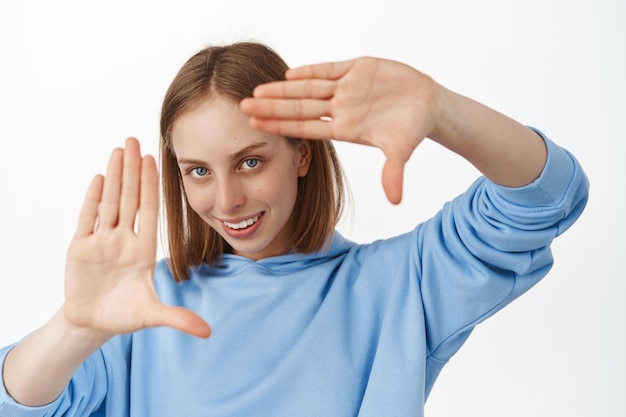 Image d'une jeune femme créative regardant à travers le geste des cadres de la main avec un sourire pensif et heureux, créant une prise de vue parfaite, recherchant à angle droit, debout sur fond blanc.