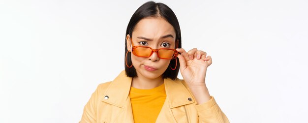 Image d'une jeune femme élégante asiatique regardant sous des lunettes de soleil avec un visage d'incrédulité sceptique exprimant un doute debout sur fond blanc Copier l'espace
