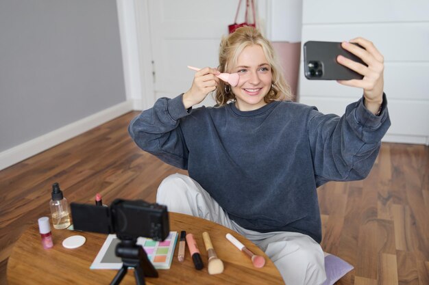 Image d'une jeune femme élégante, influente sur les réseaux sociaux, prenant des photos sur son téléphone portable en se maquillant.